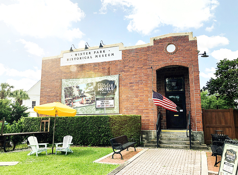 A flag flies from the Winter Park Historical Museum while a pair of Adirondack chairs sit below an umbrella in the lawn outside the museum.